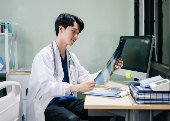 Confident young male doctor in white medical uniform sit at desk working on computer. Smiling use laptop write in medical journal in clinic.